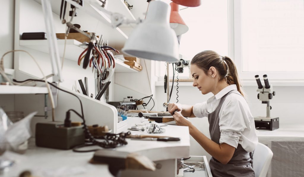 Side view of young female jeweler working on a new jewelry product at her workbench
